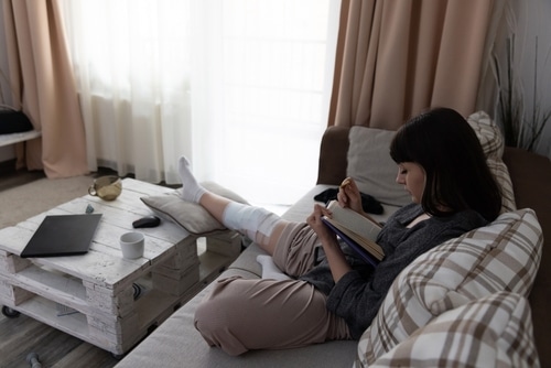 Young woman after an ACL surgery sitting on the couch and reading a book.